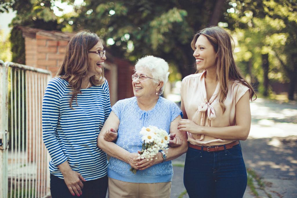 Elderly woman with flowers walking outside, accompanied by two younger women, enjoying conversation.