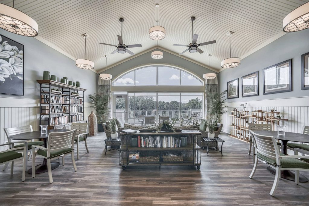 A bright, airy library with tall windows, ceiling fans, and modern furnishings. Bookshelves line the walls.
