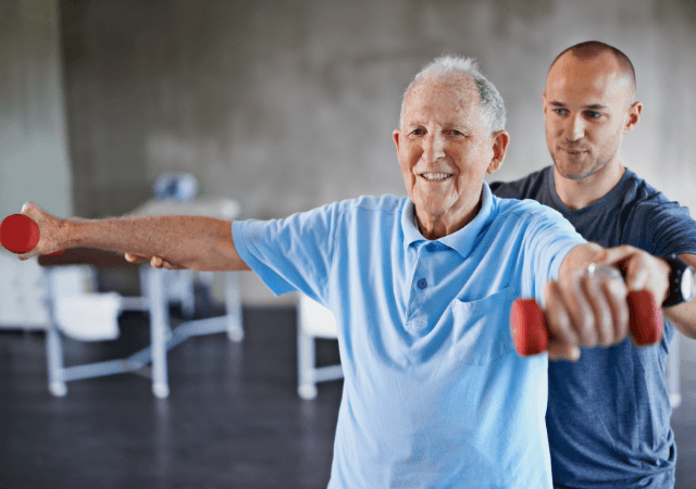 Senior man lifts hand weights with professional therapy assistant at senior living community